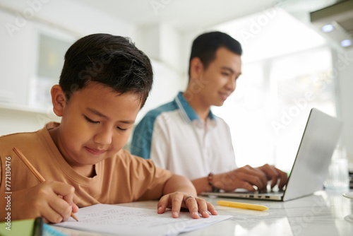 Boy doing homework when sitting next to father working on laptop at kitchen counter