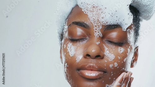 An African American woman with clear skin and closed eyes with soap suds on her face on a grey background