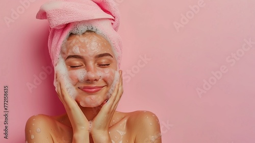 Portrait of smiling white girl washing her face with soap wearing pink towel on her head on a pink background