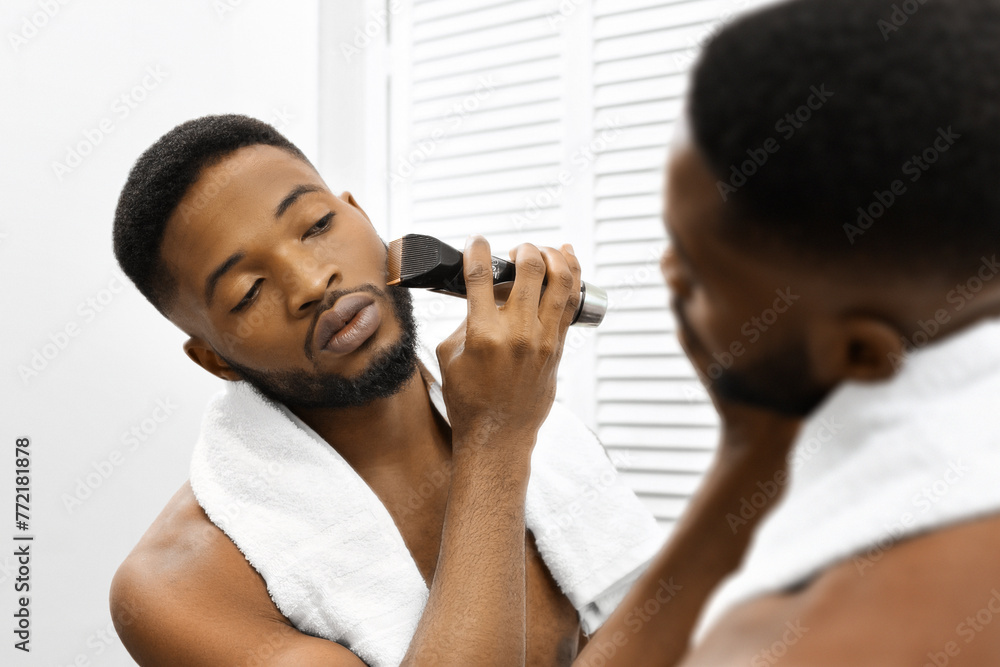 Man grooming beard with trimmer in mirror in bathroom - obrazy, fototapety, plakaty 
