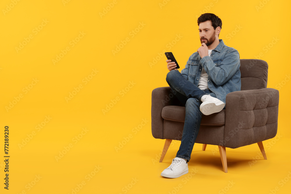 Handsome man with smartphone sitting on armchair against yellow background. Space for text