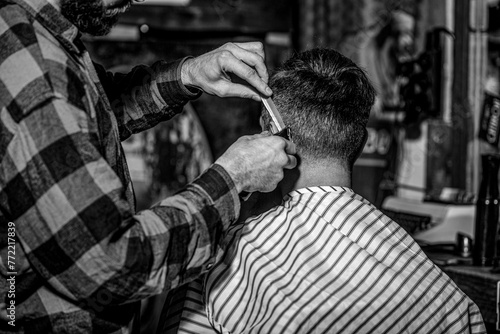 Barber guy gives a haircut to a bearded man sitting in a chair in a barbershop. Barber preparing hair clipper for bearded man, barbershop background. Barber works with a beard clipper