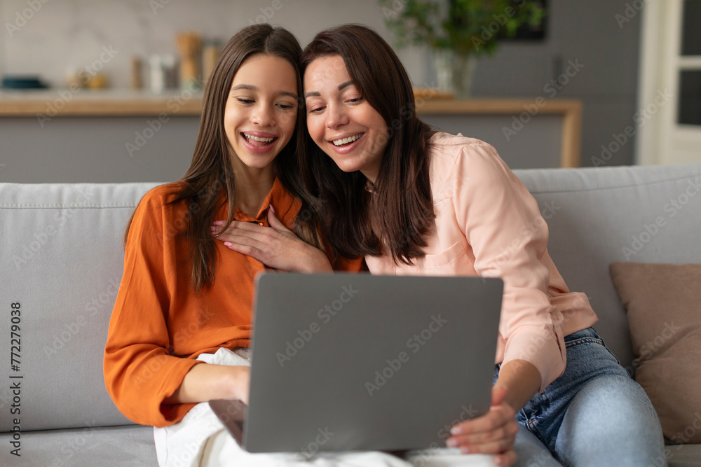 Happy mother and teen daughter looking at laptop, doing online shopping or watching movie using computer, sitting on sofa in living room Happy mother and teen daughter looking at laptop, doing online shopping or watching movie using computer, sitting on sofa in living room