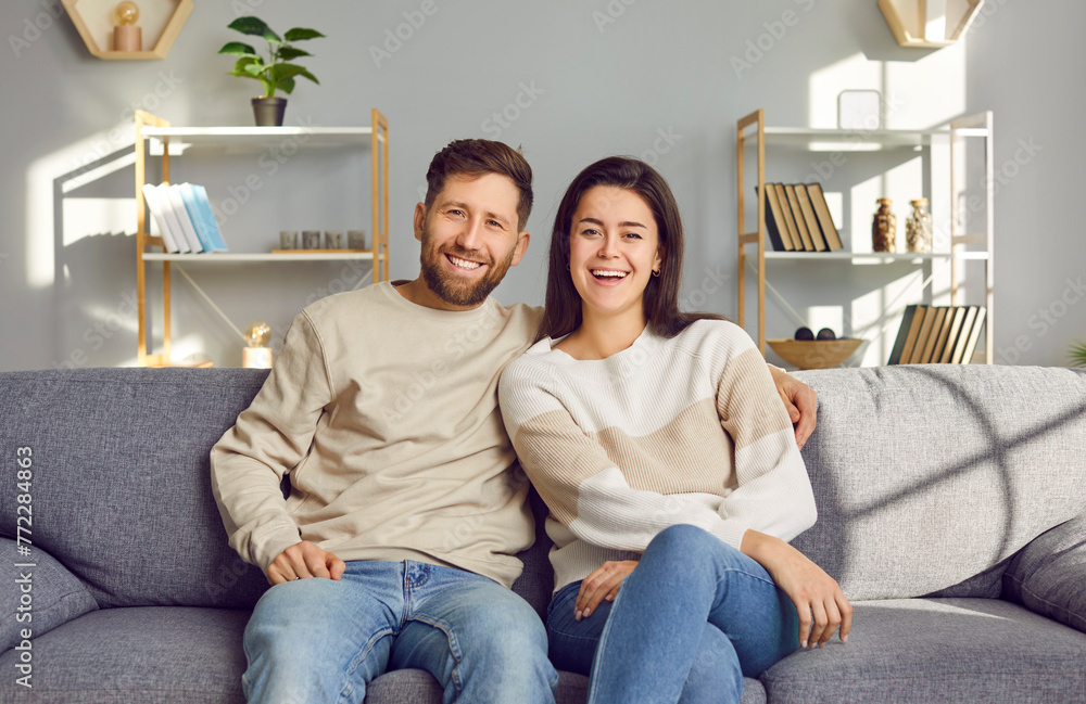 Portrait of a happy beautiful family couple sitting on the sofa at home. Cheerful young Caucasian man and woman sitting on the couch in the living room, looking at the camera and smiling