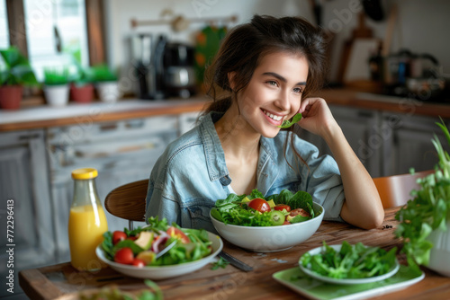 Young woman enjoying healthy food  sitting at a wooden table with green vegetables and juice bottles on it  smiling while eating salad in her home kitchen.