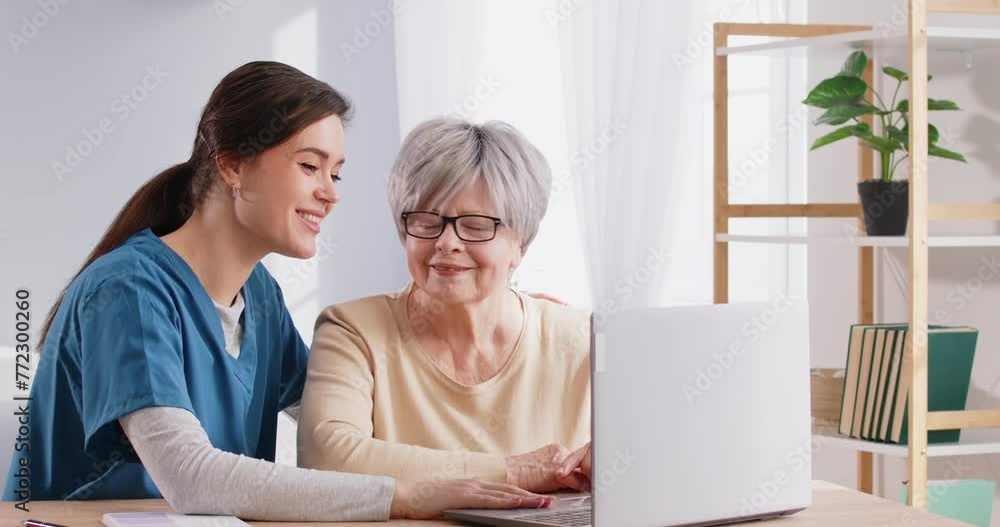 Caregiver helping retired senior woman use computer. Friendly young nurse caretaker girl showing older lady how to do web search, send emails, watch videos and open digital telemedicine app on laptop Caregiver helping retired senior woman use computer. Friendly young nurse caretaker girl showing older lady how to do web search, send emails, watch videos and open digital telemedicine app on laptop