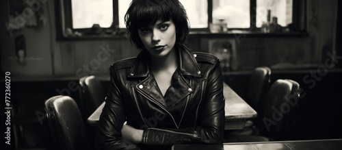A woman in a leather jacket sits at a table with her arms crossed