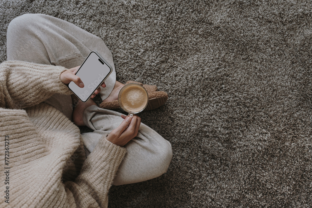 Fototapeta premium Cozy morning routine. Top view of woman wearing pastel creamy clothes sitting on fluffy grey carpet with cup of coffee and using mobile phone with blank screen. Social media mockup with copy space