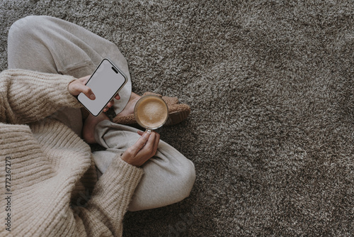 Cozy morning routine. Top view of woman wearing pastel creamy clothes sitting on fluffy grey carpet with cup of coffee and using mobile phone with blank screen. Social media mockup with copy space