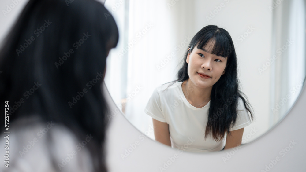 A young Asian woman gazes at her reflection in a round mirror in a bright, minimalist room. A young Asian woman gazes at her reflection in a round mirror in a bright, minimalist room.