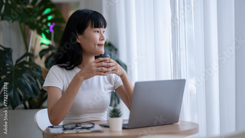 Asian woman sitting contemplating holding a coffee cup while looking away from her laptop. Looking out of the glass window In the relaxing corner of the cafe with green trees © Wasana