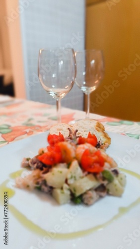 Square white plate with a starter of warm seafood salad, garnished with herbs, pachino cherry tomatoes, extra virgin olive oil and croutons, in the background two empty glasses. Mediterranean cuisine.