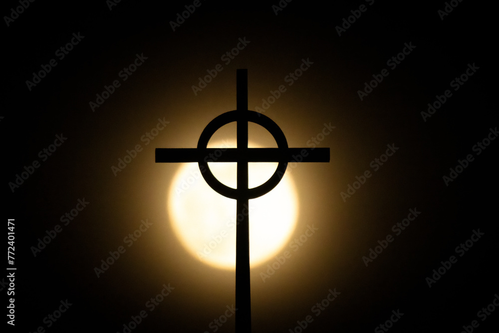 Christian Cross under spotlight. Religious symbol of the Christian faith silhouetted in front of a bright full moon, illuminating the icon of Christ. Christian Cross under spotlight. Religious symbol of the Christian faith silhouetted in front of a bright full moon, illuminating the icon of Christ.