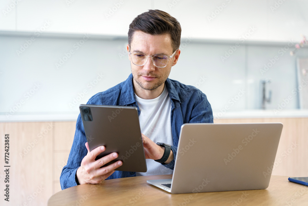 A professional man reviews content on a tablet with a laptop open before him, symbolizing modern multitasking in a workspace. A professional man reviews content on a tablet with a laptop open before him, symbolizing modern multitasking in a workspace.