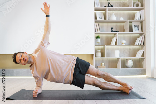 A man holds a side plank pose on a yoga mat, elevating his fitness with strength and balance, surrounded by a serene, tidy home environment.