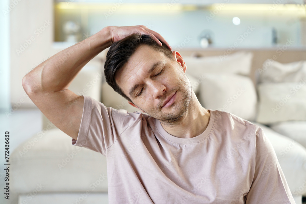 A man relaxes with a gentle neck stretch, eyes closed, enjoying a moment of tranquility in a serene and stylish living area. A man relaxes with a gentle neck stretch, eyes closed, enjoying a moment of tranquility in a serene and stylish living area.