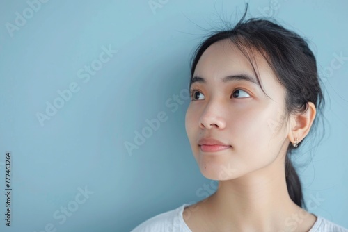 Young Asian woman with a pensive expression, gazing into the distance, blue background enhancing her serene beauty. © NS
