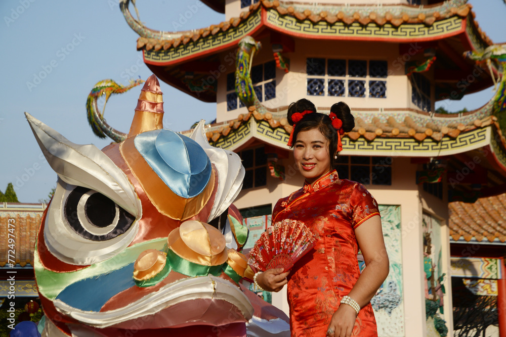 Portrait of a beautiful Asian woman in a red Chinese dress holding a fan and standing in front of a beautifully puppet lantern inside a shrine.