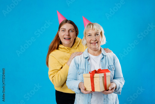 Young smiling happy daughter and mother together gifting birthday present with red ribbon isolated on blue color background studio. Human emotions, facial expression and holiday concept.