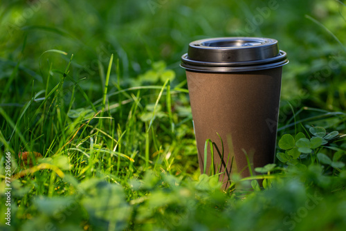 Takeaway hot coffee cup on green grass of park in summer day, meadow plants and mug in sunlight