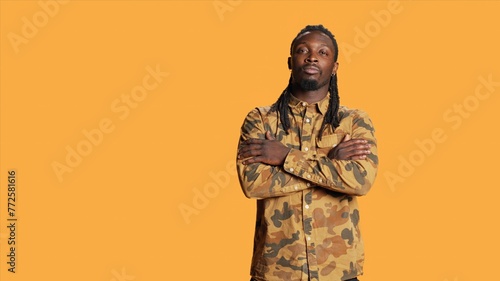Portrait of guy with dreads smiling in studio set, wearing trendy camo clothes and feeling joyful on camera. African american adult with cool hair posing over orange background, natural style.