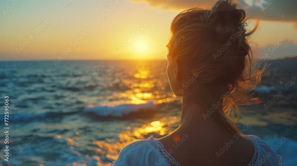 A woman is standing on the beach looking out at the ocean. The sun is setting in the background, casting a warm glow over the water. The woman is lost in thought, taking in the beauty of the scene