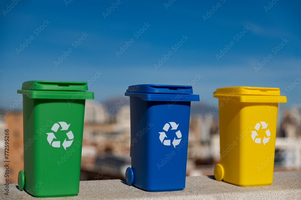 Container trash bin for recycling with city background. Yellow, green, blue bins for recycle plastic, paper and glass trash. Environmental awareness, recycling, waste management concept Container trash bin for recycling with city background. Yellow, green, blue bins for recycle plastic, paper and glass trash. Environmental awareness, recycling, waste management concept