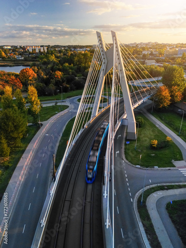 Drone forward overhead view of suspension bridge with tram movement in Krakow, Poland. Sunset top view of bridge structure and public transport in motion blur, empty asphalt road and park below photo