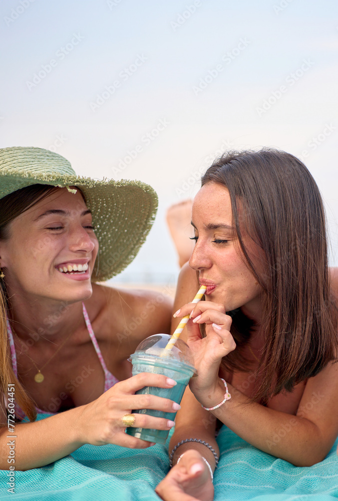 Vertical. Two attractive Caucasian women smiling and holding and drinking natural smoothies in summer holidays. Beautiful female friends enjoy vacation lying on the beach