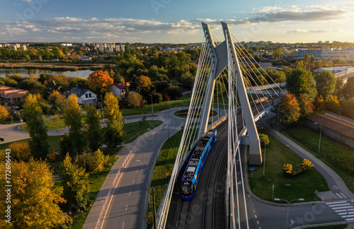 Aerial perspective view of city road and modern bridge in Krakow, Poland. Drone forward above view of tram driving on circular steel and concrete bridge construction, autumn park in sunset rays