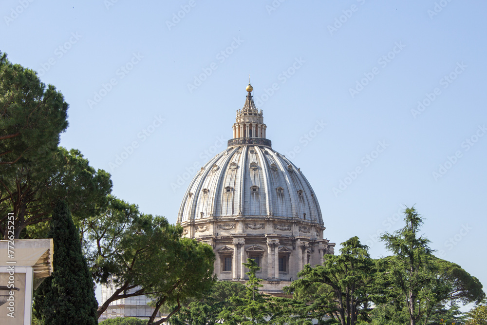 Fototapeta premium View of St. Peter's Basilica's iconic dome rising majestically above green trees under a clear blue sky, a symbol of architectural brilliance
