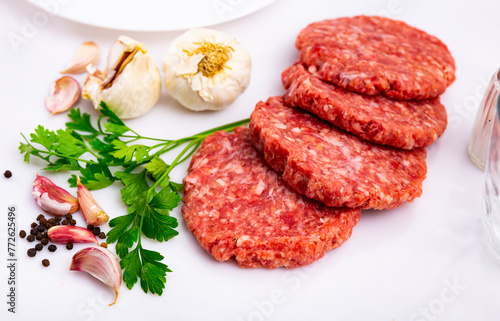 Raw pork patty burgers if form of ground meat on a plate decorated with peppercorns, garlic and parsley on white background