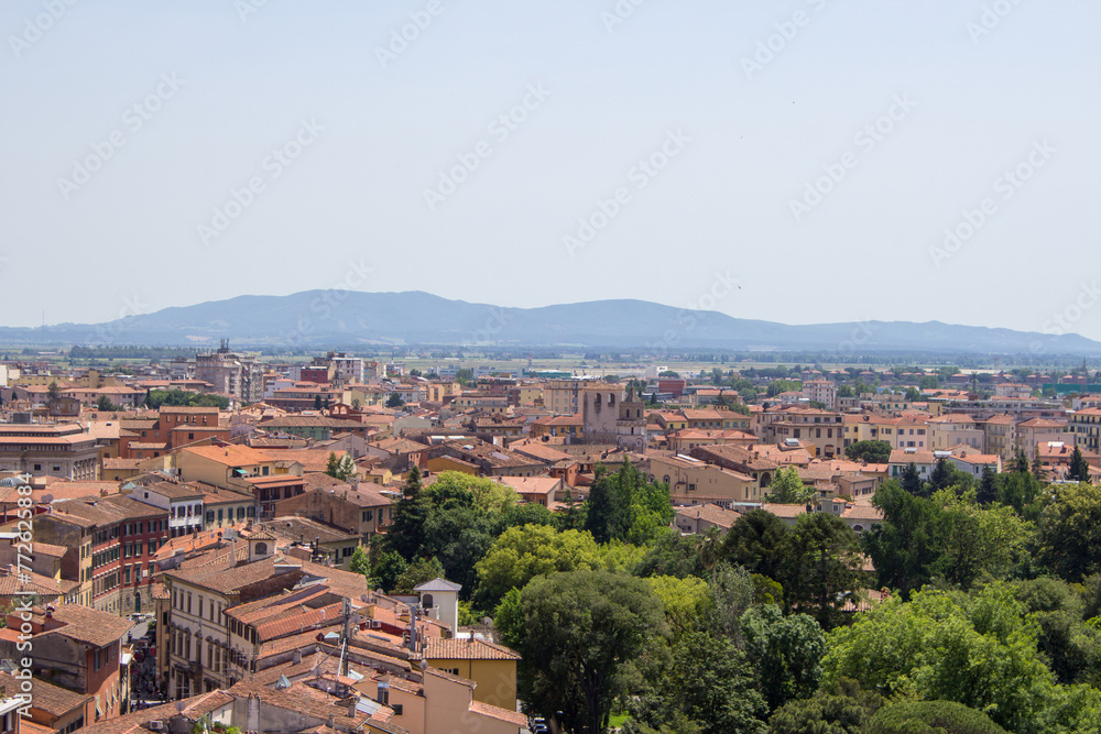 The dense urban landscape of Pisa unfolds beneath a hazy sky, with the distant Tuscan hills framing the historic city.
