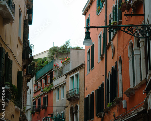 A narrow alley in Venice showcases vibrant living spaces with flourishing balcony gardens, ornate window shutters, and traditional street lamps.