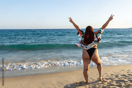 Plus size Latina woman looking at the sea making the peace symbol with her hands. Young black girl with braids enjoying a day at the beach on her summer vacation. photo