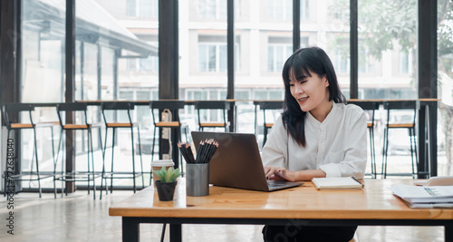 Smiling Asian woman focused on her laptop work in a bright cafe environment with coffee and stationery on the table.