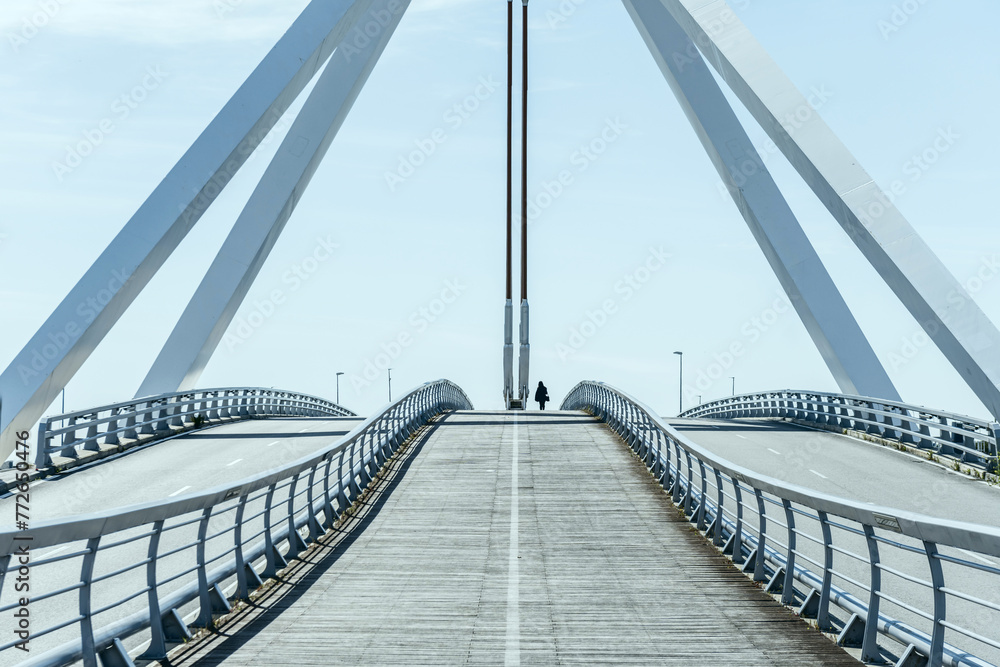 Metal structure of a small bridge with hanging metal structure, walkways and wooden floors for pedestrians on a nice sunny day with a lone pedestrian crossing it