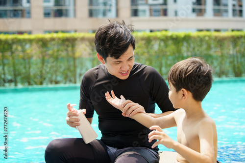 Father is applying a sun screen or sun block lotion on his son body before going to swimming in the swimming pool. Weekend vacation activities between father and son. photo