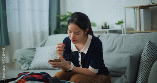 Selective focus of Asian teenager woman sitting on sofa making checklist of things to pack for travel, Preparation travel suitcase at home.