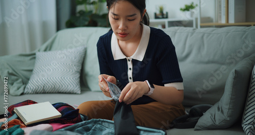 Front view of Asian teenager woman sitting on sofa packing travel luggage with personal items for traveling trip, Preparation travel suitcase at home.