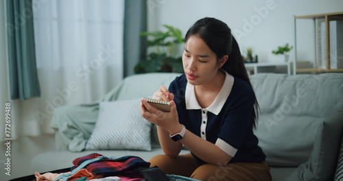 Selective focus of Asian teenager woman sitting on sofa making checklist of things to pack for travel, Preparation travel suitcase at home.