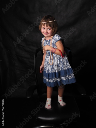 little girl in a blue dress and jewelry on a black background in the studio in full growth (ID: 772729867)