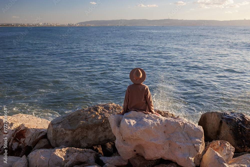 Back view. relaxed girl in a hat and coat sitting in front of the sea on the stones. Back view. relaxed girl in a hat and coat sitting in front of the sea on the stones.