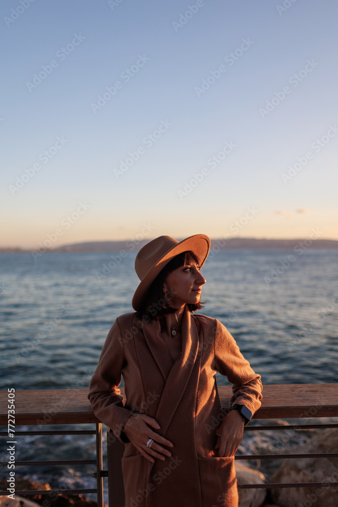 Portrait of a charming young woman in a coat and hat looking at the sea. A girl stands on the embankment and looks at the sea.