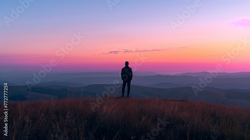A person stands as a silhouette atop a mountain peak  overlooking a vibrant sunset that paints the sky with hues of red and blue. 