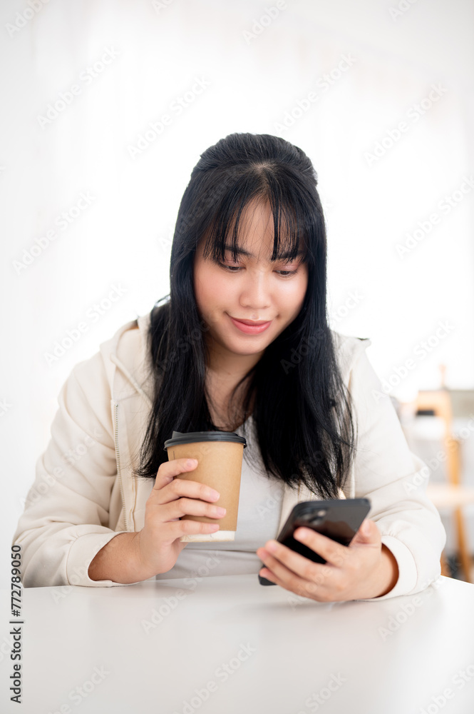A positive, smiling Asian woman in casual wear is sipping coffee and using her smartphone in a cafe. A positive, smiling Asian woman in casual wear is sipping coffee and using her smartphone in a cafe.