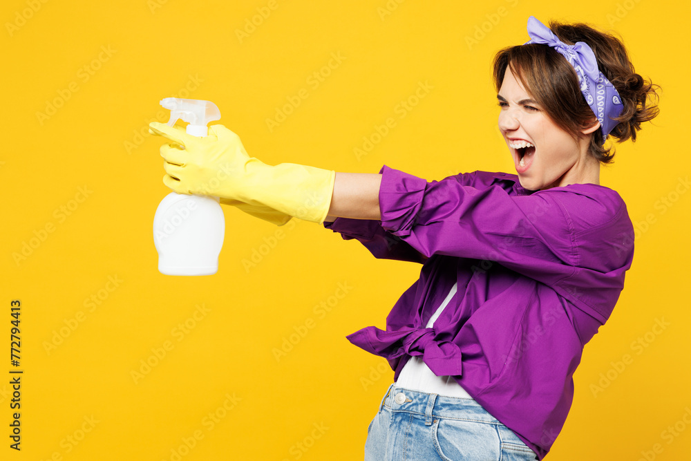 Side profile view excited young woman wears purple shirt rubber gloves do housework tidy up hold in hand use spray bottle isolated on plain yellow background studio portrait. Housekeeping concept. Side profile view excited young woman wears purple shirt rubber gloves do housework tidy up hold in hand use spray bottle isolated on plain yellow background studio portrait. Housekeeping concept.