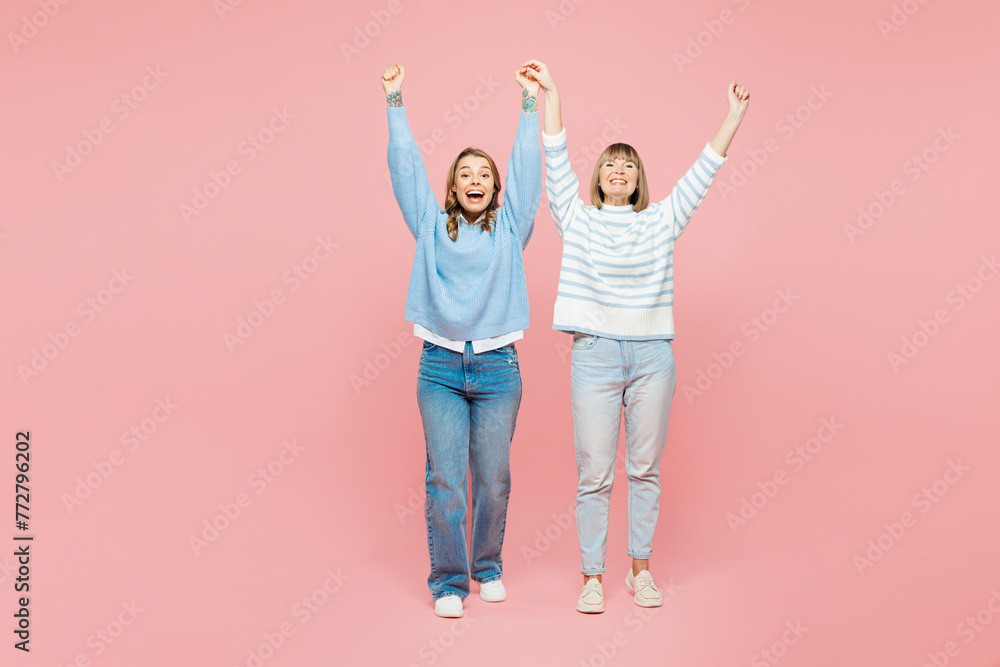 Full body elder parent mom with young adult daughter two women together wears blue casual clothes raise up hands do winner gesture isolated on plain pastel light pink background. Family day concept. Full body elder parent mom with young adult daughter two women together wears blue casual clothes raise up hands do winner gesture isolated on plain pastel light pink background. Family day concept.