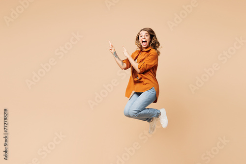 Full body side view young fun woman she wear orange shirt casual clothes jump high point index finger aside on area isolated on plain pastel light beige background studio portrait. Lifestyle concept.