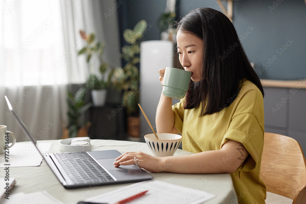 Medium shot of young Asian university student sitting at dining table at home drinking coffee and using laptop Medium shot of young Asian university student sitting at dining table at home drinking coffee and using laptop
